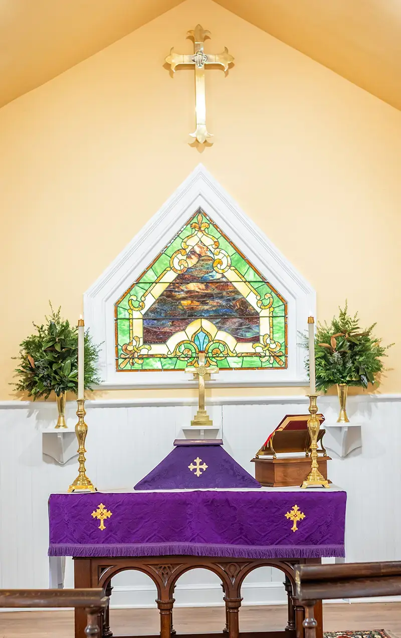 Church altar with a purple cloth, cross, candlesticks, and stained glass above.