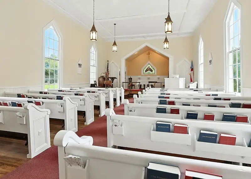 Sunlit church interior with white pews, tall windows, and a central aisle, highlighting the space available for gatherings and events.