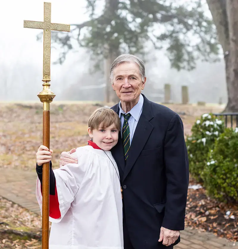 A young acolyte in white vestments holds a processional cross alongside an adult parishioner on a misty morning