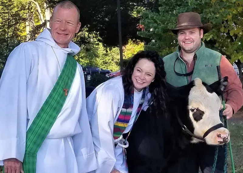 Members of the church leadership smile outdoors beside a black-and-white cow, reflecting community life and pastoral presence.