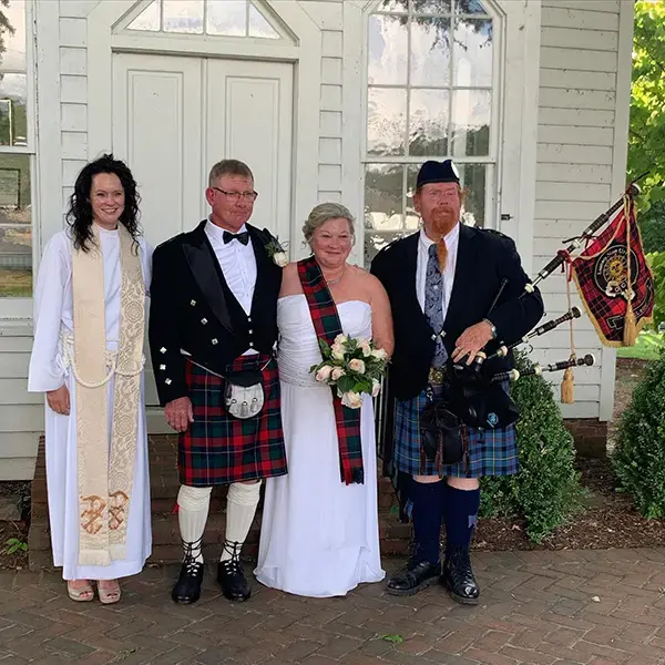 A wedding party poses outside the church with the officiant, bride, groom in tartan kilt, and bagpiper