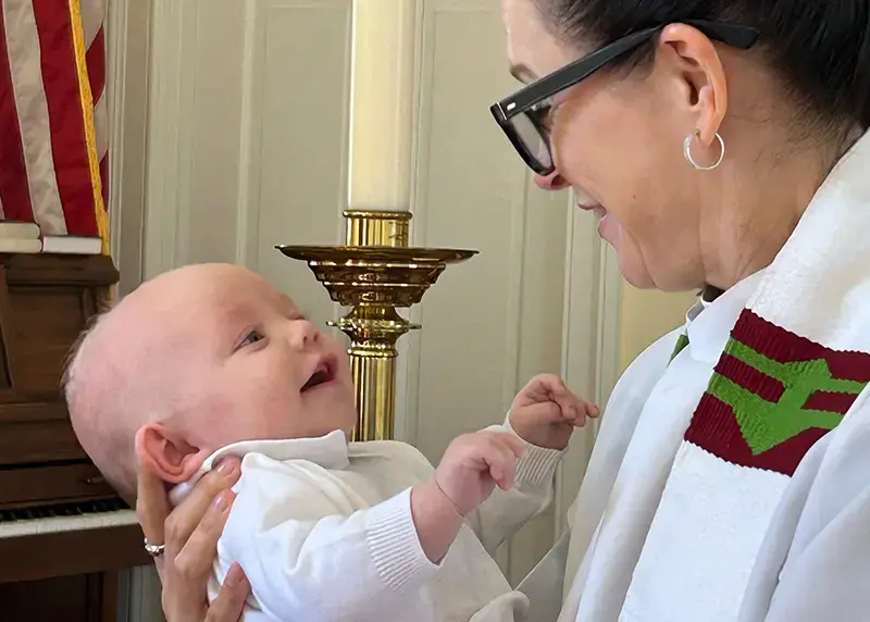 A priest holds an infant during a baptism ceremony with the paschal candle in the background